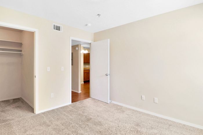 Bright and inviting bedroom with plush carpet and open closet, complemented by a view of a cozy hallway leading to a warm, wood-finished kitchen.