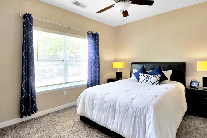 Inviting bedroom with plush bedding, modern decor, and ample natural light from a large window dressed with elegant drapes.