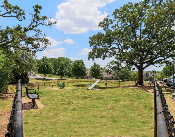 Charming community park with lush greenery, benches, playground equipment, and mature trees under a bright blue sky.