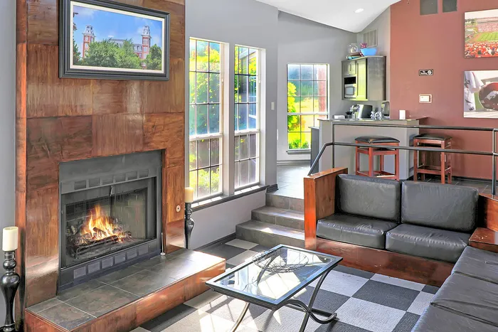 Charming living room with a cozy fireplace, modern furnishings, and abundant natural light from large windows overlooking greenery.