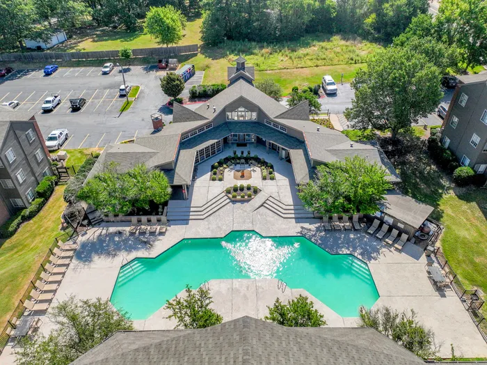 Aerial view of an upscale residential complex featuring a uniquely shaped turquoise pool, surrounded by modern architecture, lush greenery, and ample parking.