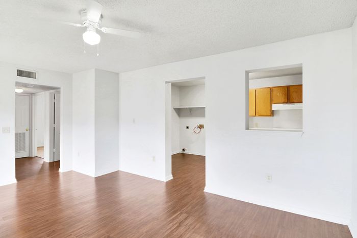 Bright and airy apartment interior featuring sleek wood flooring, a ceiling fan, and a passthrough kitchen window. Modern and inviting living space with ample natural light.