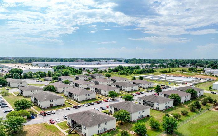 Aerial view of a spacious apartment community with neatly arranged buildings, surrounded by lush greenery under a bright blue sky.