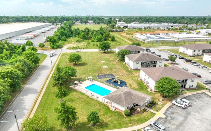 Charming aerial view of an inviting residential complex with well-maintained apartments, lush greenery, a sparkling blue swimming pool, and a fun playground, set amidst a serene suburban area.