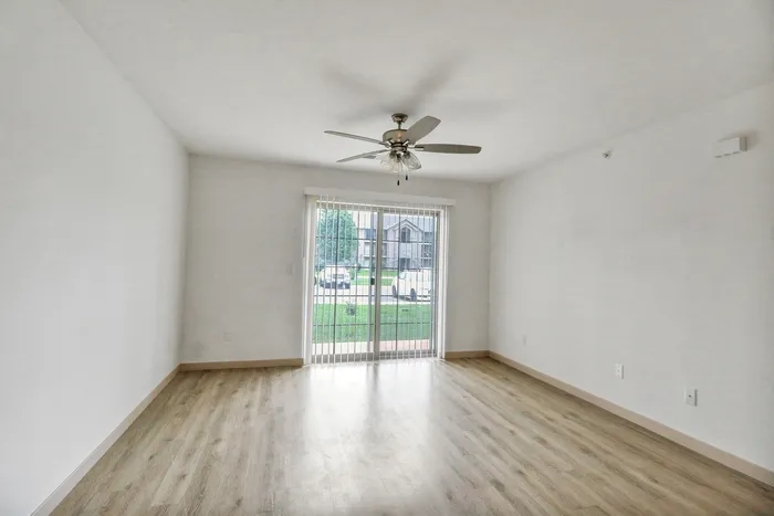 Bright and airy living room with elegant wooden flooring, large sliding glass doors, and a modern ceiling fan, offering a serene view of the green outdoors.