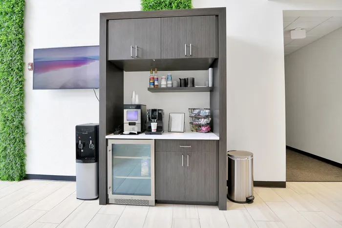 Modern refreshment station with sleek cabinetry, built-in beverage fridge, coffee machine, and stylish shelving against a bright wall with green accents.