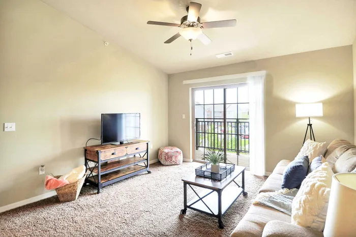 Bright and airy living room with plush carpet, cozy sofa, and elegant decor. Features a ceiling fan, large TV, and expansive sliding doors leading to a sunlit balcony.