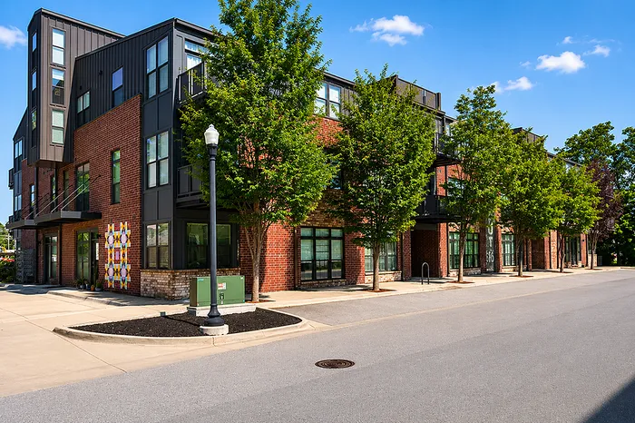 Modern apartment complex with stylish brick facade, large windows, and lush greenery lining the street under a clear blue sky.