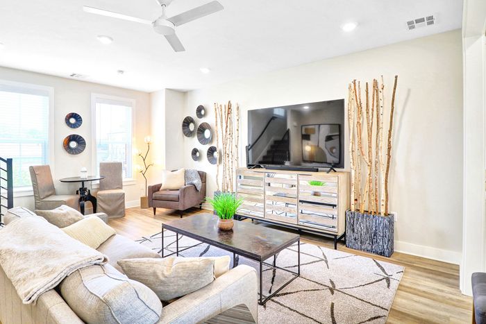 Bright and inviting living room with stylish decor, featuring a comfortable sofa, elegant chairs, and a large flat-screen TV. Natural light fills the room through large windows, complementing the modern wooden accents and lush indoor plant.