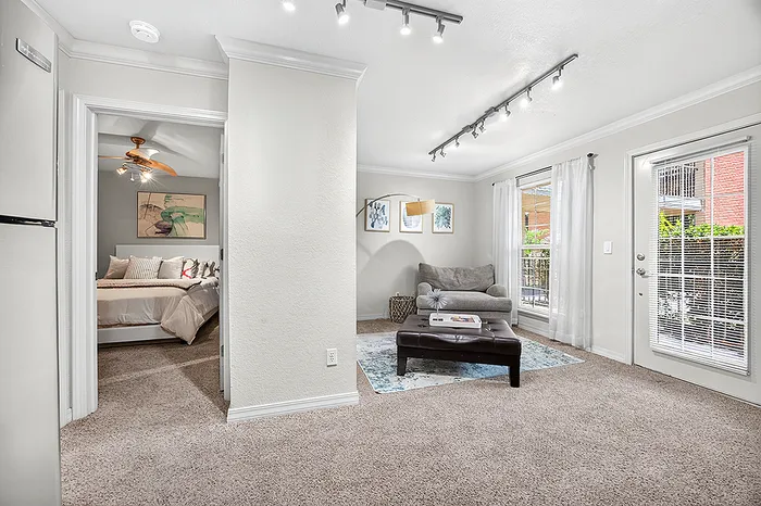 Bright and inviting living area with plush carpeting, cozy seating, and an abundance of natural light from large windows; adjacent view into a welcoming bedroom with elegant decor.
