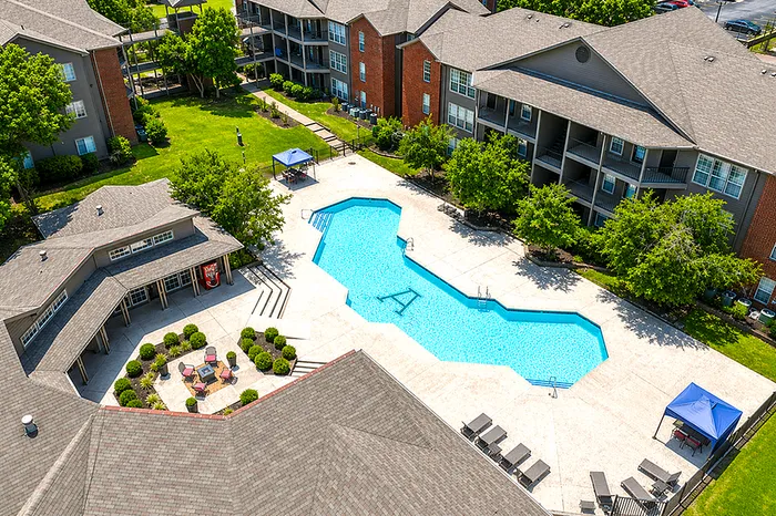 Aerial view of a luxurious apartment complex with a shimmering pool, surrounded by modern buildings and lush greenery.