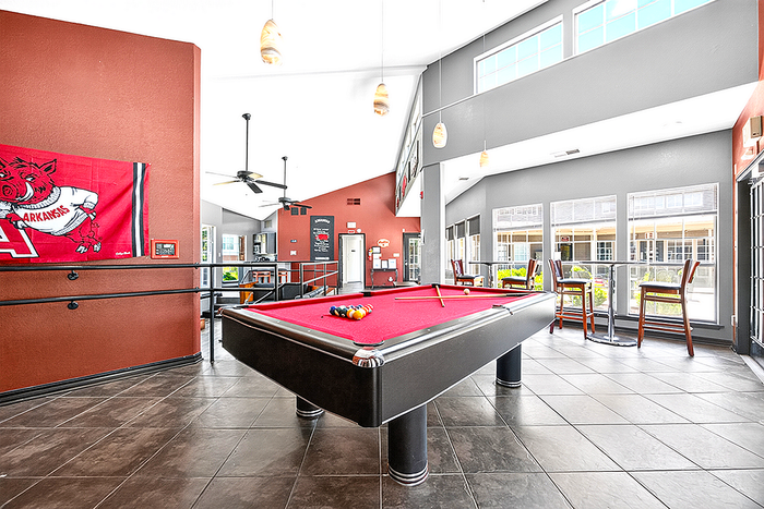 Modern apartment lounge with a vibrant red pool table, large windows, high ceilings, and cozy seating overlooking a sunlit courtyard.