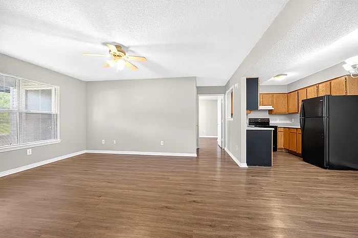 Spacious open-concept living area with sleek hardwood floors, modern ceiling fan, and natural light from large windows, adjacent to a kitchen with stylish wooden cabinetry.
