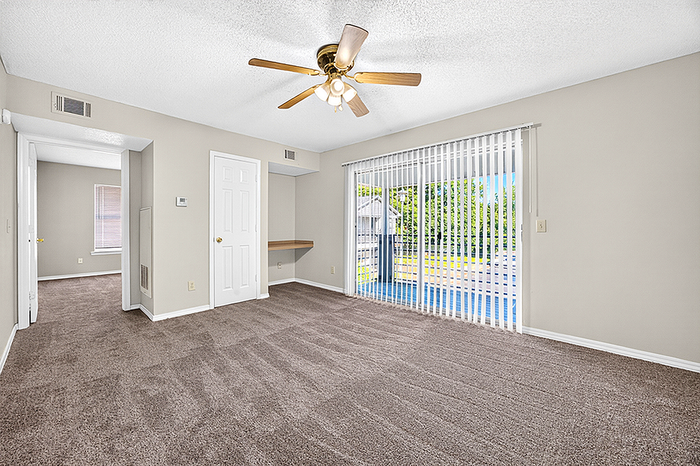 Bright and spacious living room featuring plush carpeting, large windows with vertical blinds, and an elegant ceiling fan, perfect for relaxation and comfort.
