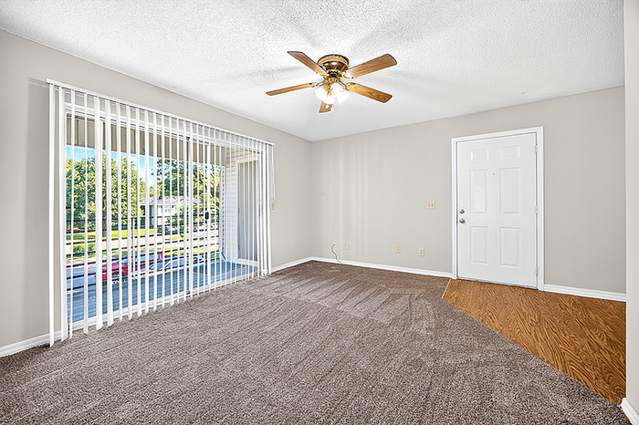Bright and spacious living room with plush carpeting, ceiling fan, and floor-to-ceiling windows offering a beautiful outdoor view, perfect for relaxation.