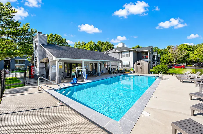 Inviting community pool area with lounge chairs and a clubhouse under a clear blue sky, surrounded by lush greenery and modern apartments.
