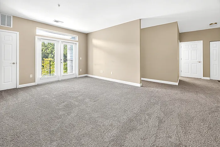 Bright and spacious living area with plush carpeting, large windows, and elegant double doors leading to a sunny balcony.