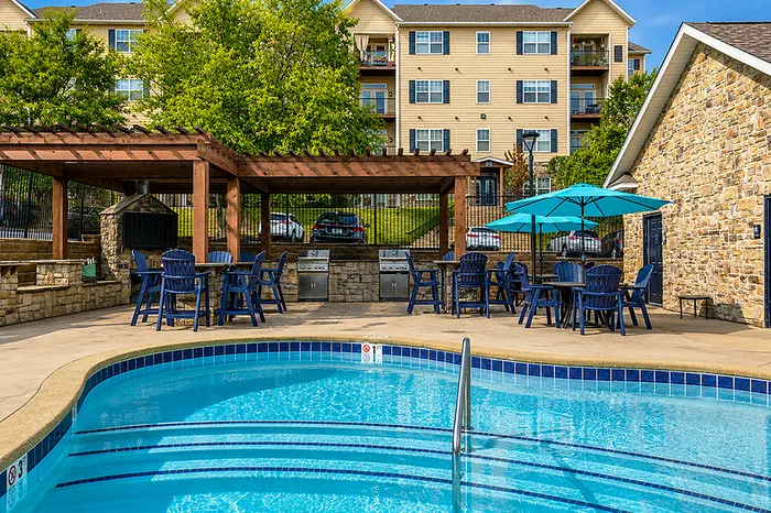 Inviting apartment pool area with stylish blue lounge chairs, shaded seating under wooden pergolas, and a BBQ grill station set against a backdrop of charming residential buildings.