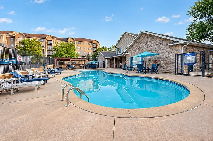 Inviting residential community with a sparkling pool, stylish stone clubhouse, and comfortable lounge chairs set against a vibrant backdrop of trees and modern apartments.