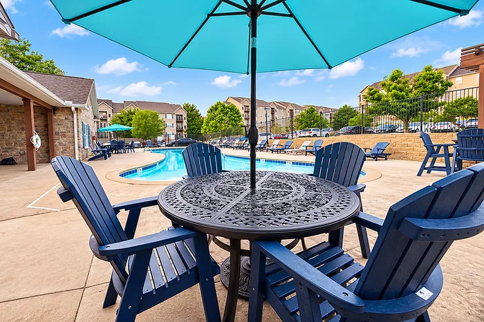 Charming outdoor pool area with elegant patio furniture under a bright blue umbrella, surrounded by lush greenery and modern apartment buildings.
