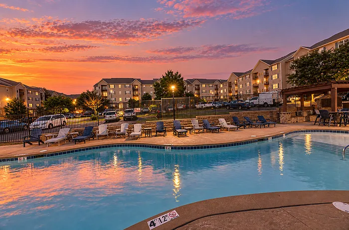 Serene apartment complex featuring a sparkling pool and lounging chairs under a vibrant sunset sky, perfect for relaxation and leisure.