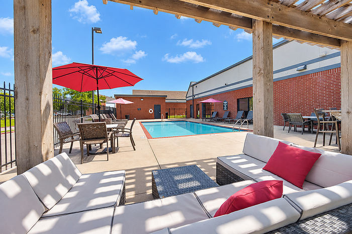 Charming outdoor pool area with shaded seating, vibrant red umbrellas, and brick building backdrop under a bright blue sky.