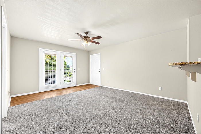 Bright and airy living room with plush carpeting, ceiling fan, and large windows leading to a charming balcony.