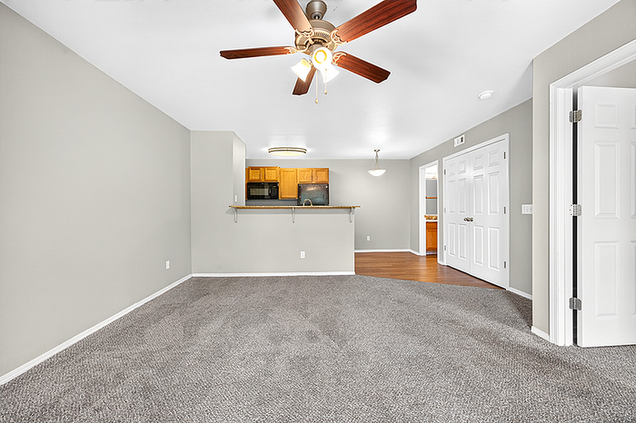 Inviting open-plan living room with plush carpet, ceiling fan, and modern kitchen in the background.