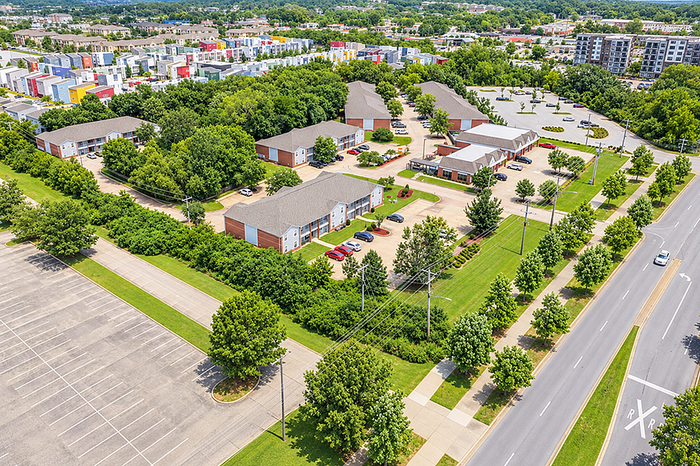 Aerial view of a serene apartment complex surrounded by lush green trees, showcasing modern buildings with ample parking, well-maintained pathways, and vibrant community spaces.