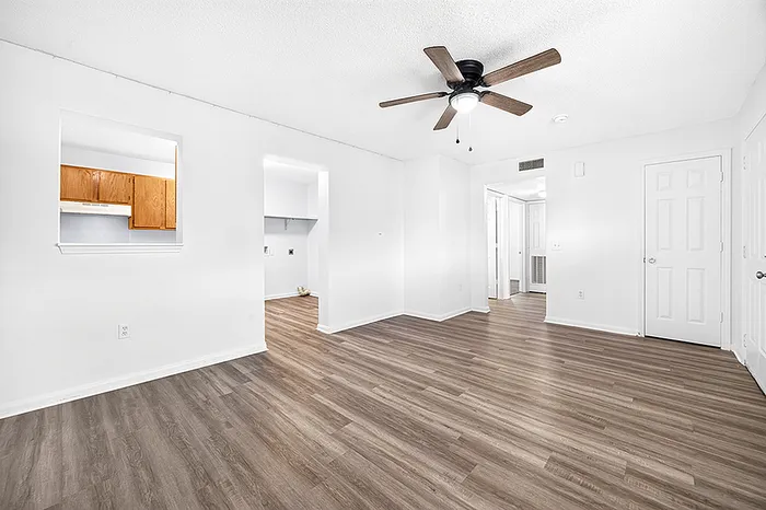 Spacious living room with sleek wood flooring and modern ceiling fan, featuring a glimpse into a bright kitchen space with wooden cabinets.