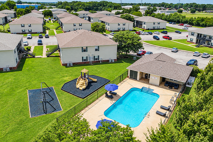 Aerial view of a vibrant apartment complex featuring multiple buildings surrounded by lush greenery, a central playground, and a sparkling blue swimming pool with a relaxation area.