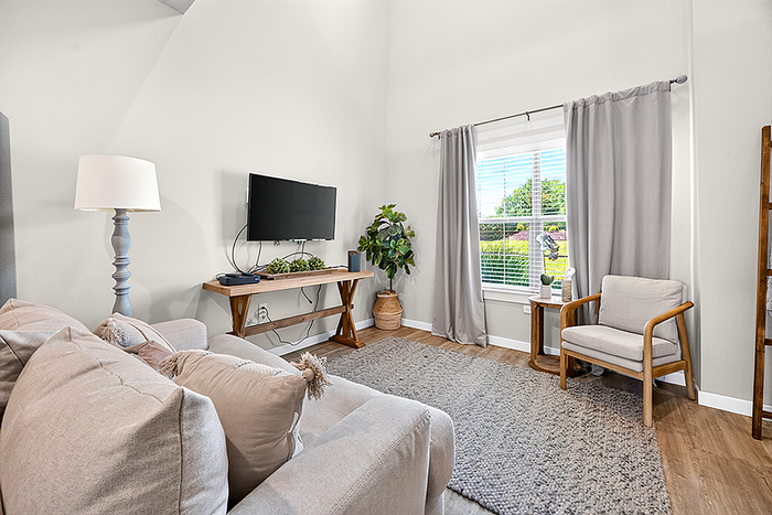Charming living room with cozy beige sofas, flat-screen TV, soft grey curtains, and abundant natural light from a large window, creating a warm and inviting atmosphere.