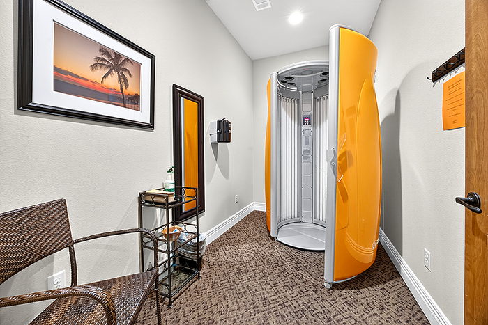 Chic tanning room featuring modern vertical tanning booth, stylish wicker chair, and soothing beach artwork.