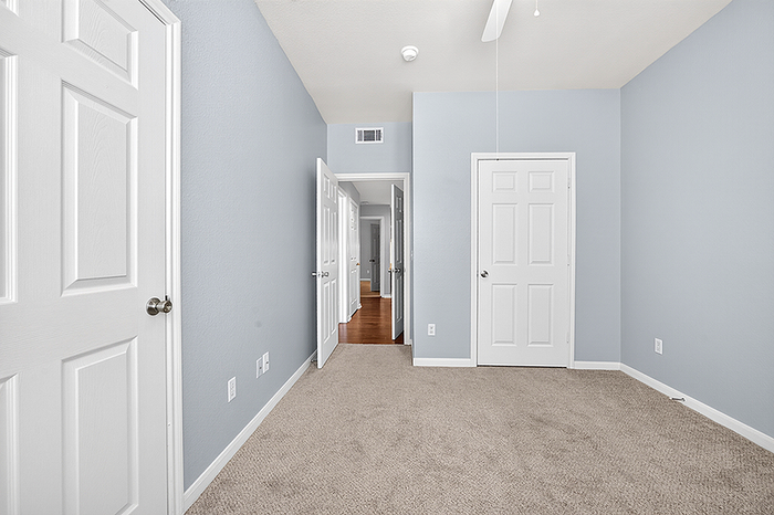Bright and inviting bedroom with soft blue walls, plush carpeting, and ample natural light. Features multiple doors for easy access and a modern ceiling fan for comfort.