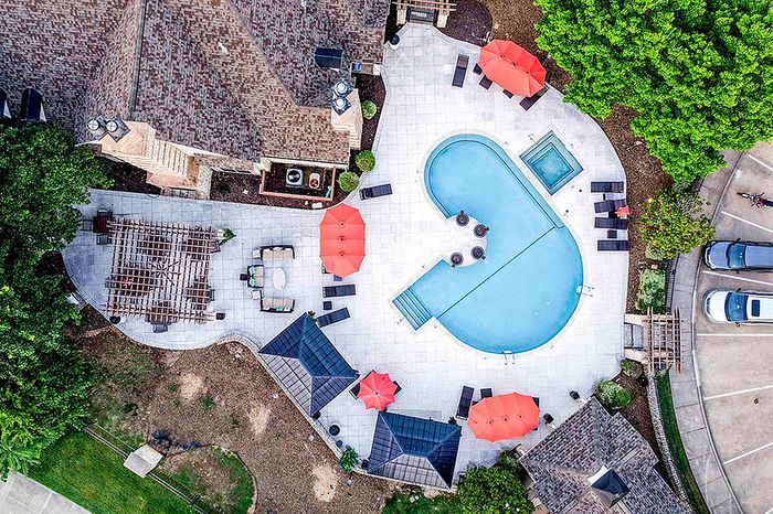 Vibrant aerial view of luxurious apartment complex with a uniquely shaped swimming pool, inviting sun loungers, and stylish red umbrellas, surrounded by lush greenery.