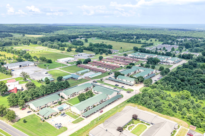 Aerial view of a charming residential community surrounded by lush greenery, showcasing multiple apartment buildings with green rooftops and ample parking, set in a serene and spacious environment.
