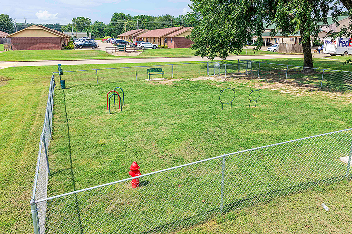 Charming fenced dog park with lush green grass, playful structures, and cozy seating within a welcoming community setting.