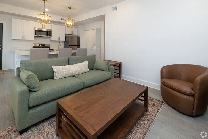 Inviting living area with a modern green sofa, elegant wood coffee table, plush brown armchair, and a sleek kitchen with stylish pendant lights in the background.