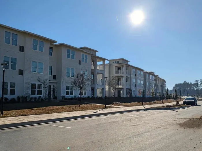Modern apartment complex with sunlight reflecting off contemporary white buildings, surrounded by scenic greenery and clear blue skies.