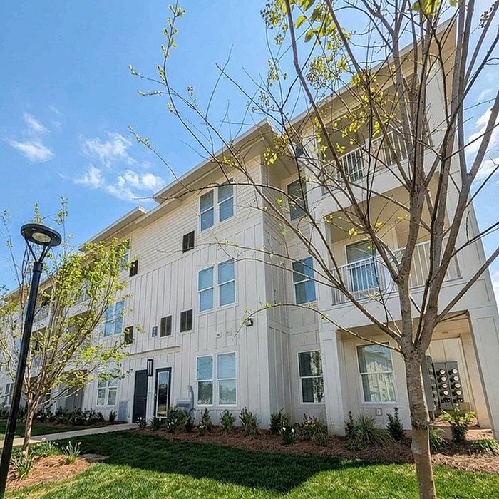 Charming three-story apartment building with modern architecture surrounded by lush greenery and a bright blue sky.