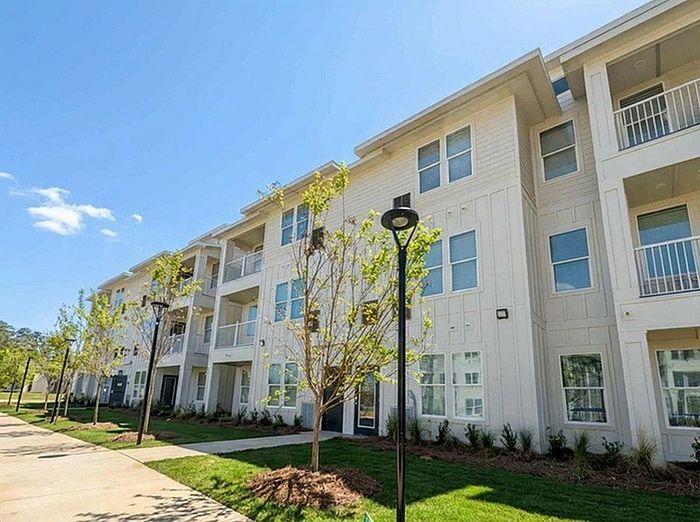 Charming three-story apartment building with modern design, surrounded by lush greenery and a clear blue sky.