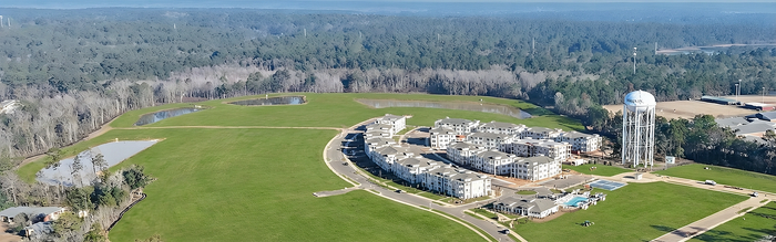 Scenic aerial view of modern apartment complex surrounded by lush greenery and serene landscape, featuring a water tower and nearby ponds.