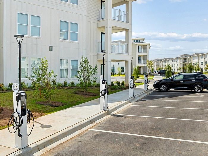 Modern apartment building with electric car charging stations and ample parking space, surrounded by lush greenery.