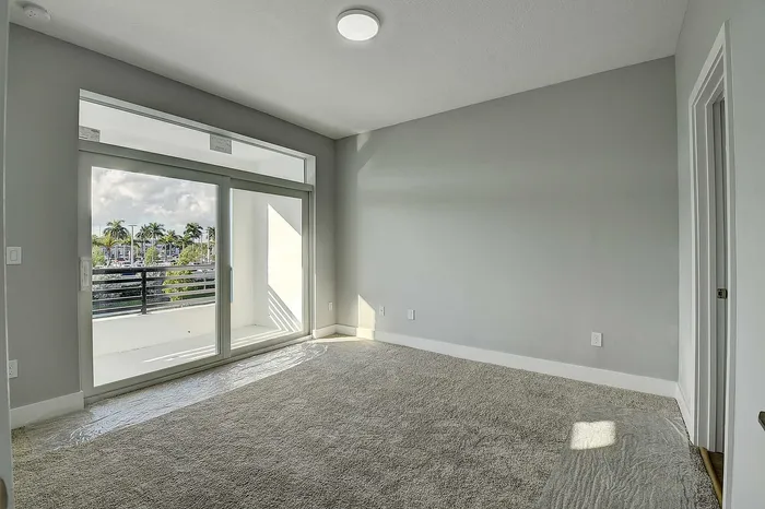 Bright, spacious room with plush carpet and large glass doors opening to a balcony view of lush palm trees.