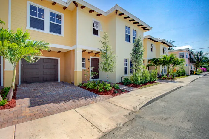 Charming townhouse with a sunny yellow exterior, lush landscaping, and a cozy front porch. Ideal location with palm trees lining the street.