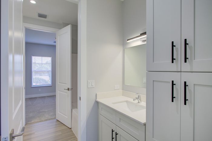 Bright and modern bathroom with sleek cabinetry and a spacious adjoining bedroom featuring soft carpet and a large window.