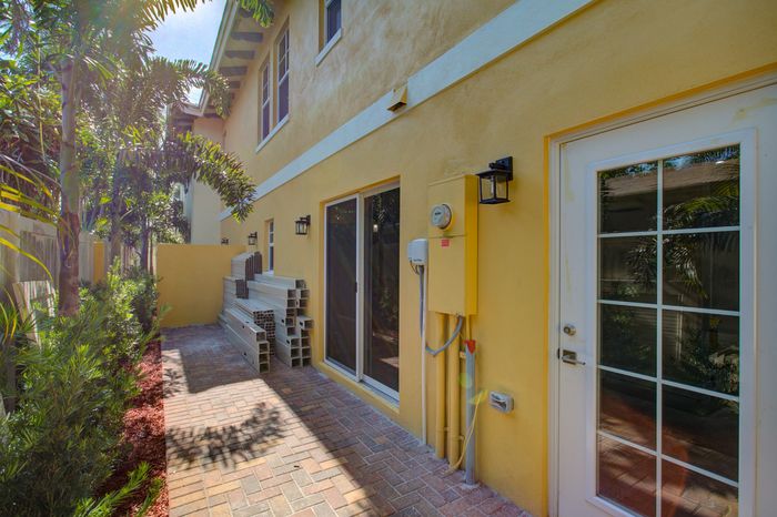Charming exterior side view of a sunny yellow building with elegant lantern lighting and lush tropical plants on a paved path.