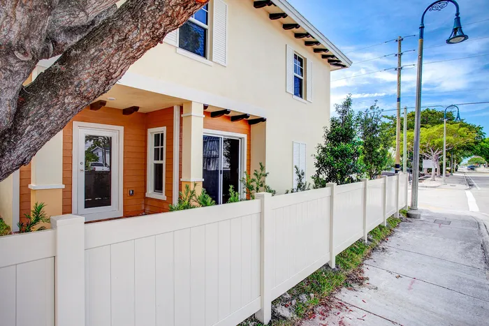 Charming two-story home with a warm peach and cream facade, nestled behind a pristine white fence, surrounded by lush greenery and shaded by a large tree, creating an inviting and serene street view.
