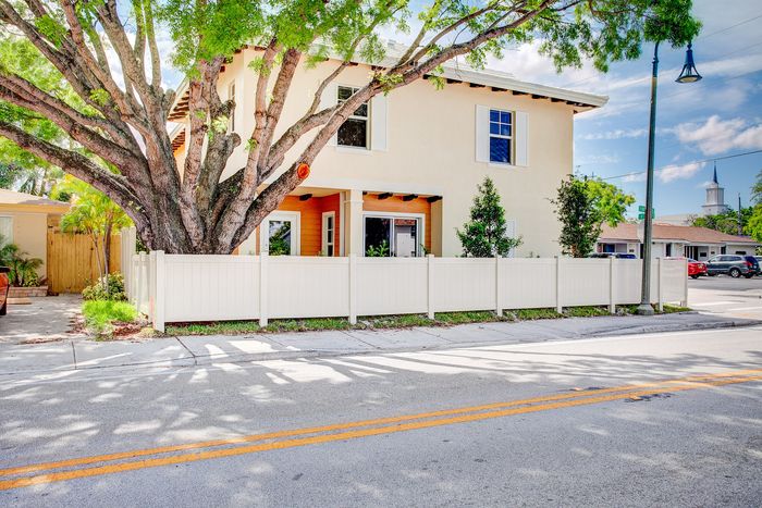 Charming two-story home with a white picket fence and lush greenery, nestled under a large, majestic tree in a peaceful neighborhood.