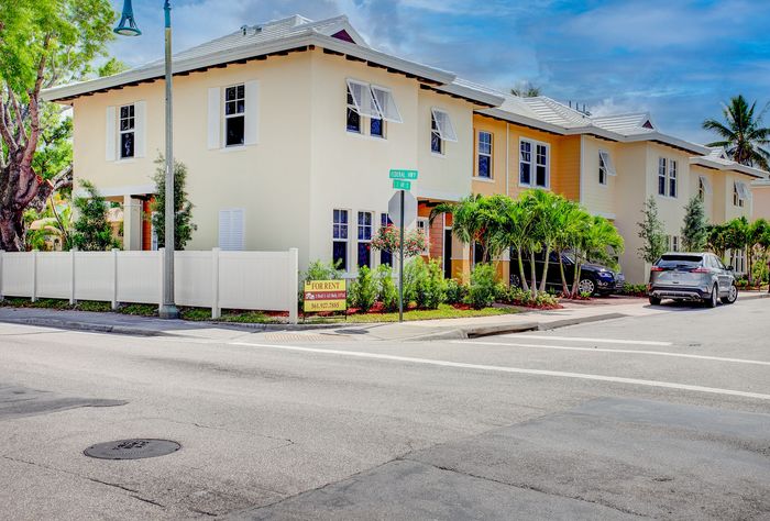 Charming corner townhouse with a sunny facade, surrounded by lush greenery and a welcoming white fence, perfect for rental opportunities.